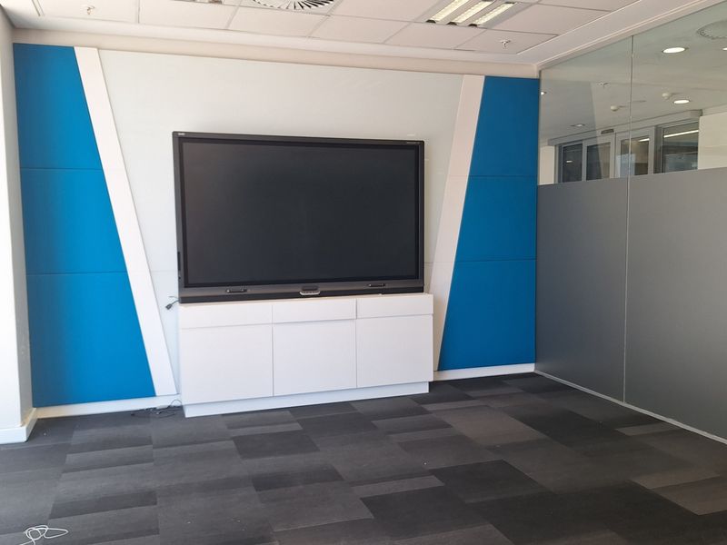 A modern office meeting room featuring acoustic wall paneling, a built-in media cabinet, glass partitioning, and professional carpet tiles.
