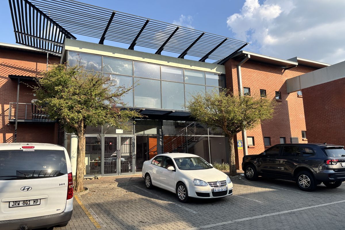 Front elevation of an industrial building featuring a brick facade, large glass curtain wall, and a modern metal louvered roof structure with paved parking.