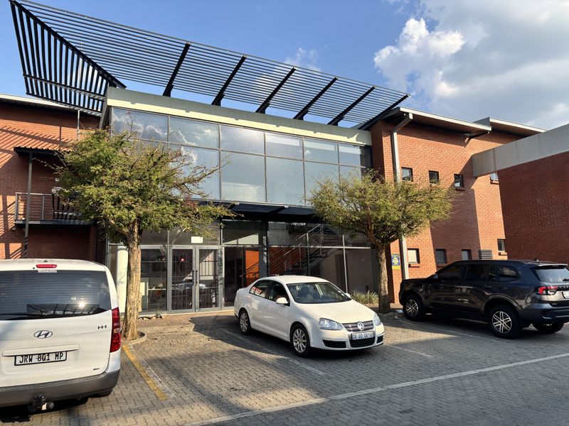 Front elevation of an industrial building featuring a brick facade, large glass curtain wall, and a modern metal louvered roof structure with paved parking.
