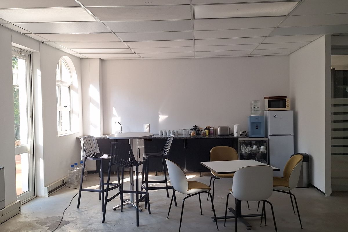 A functional office kitchenette featuring dark cabinetry, a sink, and a drop ceiling, with an adjacent dining area and arched window.