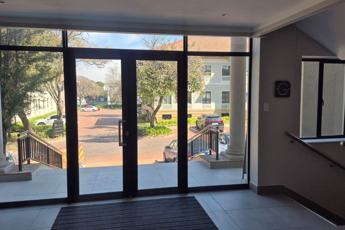 Bright and modern office building entrance featuring large glass double doors, tiled flooring, and a staircase with a sleek metal handrail.