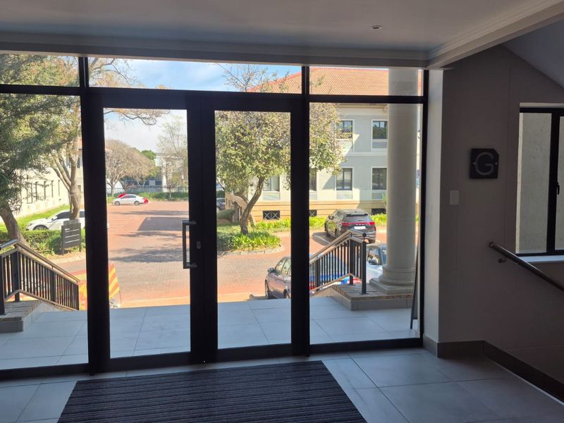 Bright and modern office building entrance featuring large glass double doors, tiled flooring, and a staircase with a sleek metal handrail.
