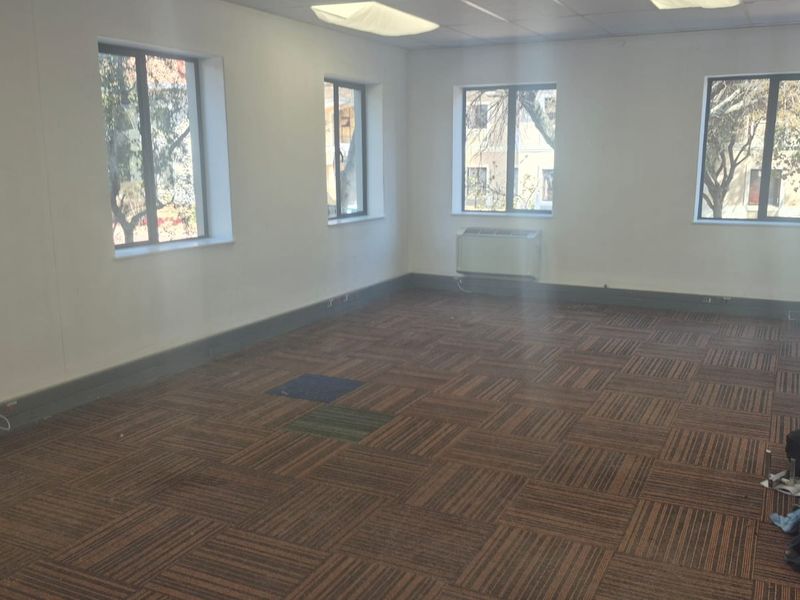 A spacious, empty office area featuring carpet tile flooring, suspended ceilings with recessed lighting, and multiple windows providing natural light.