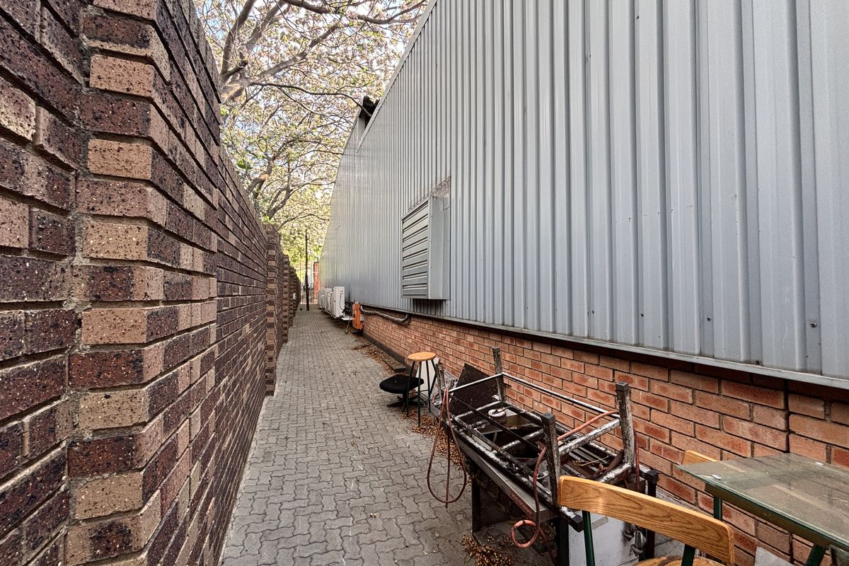 A narrow paved side passage between a brick boundary wall and the corrugated metal exterior wall of an industrial building.
