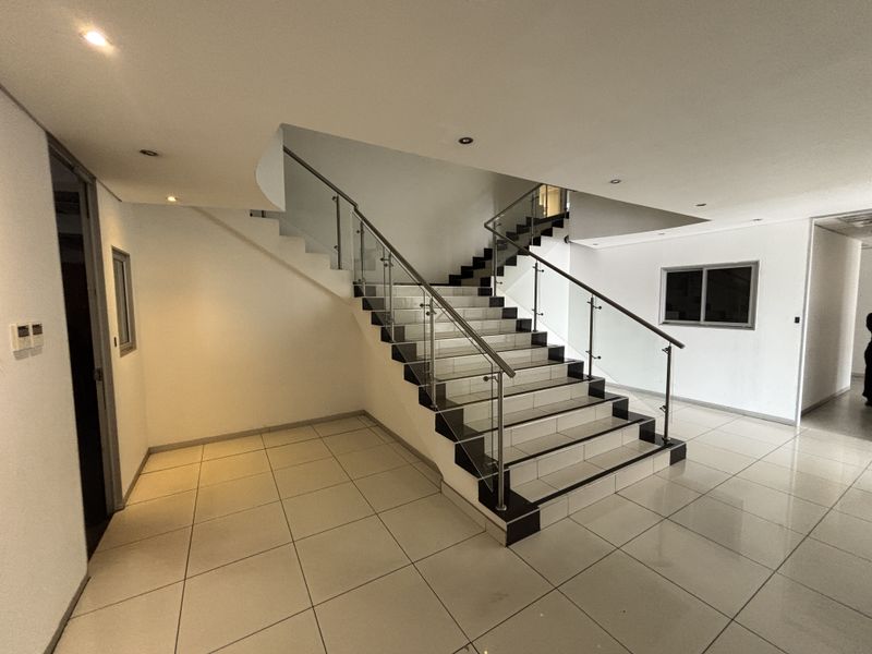 Spacious industrial hallway featuring a modern tiled staircase with a glass balustrade and stainless steel handrail, complemented by recessed lighting and polished floor tiles.