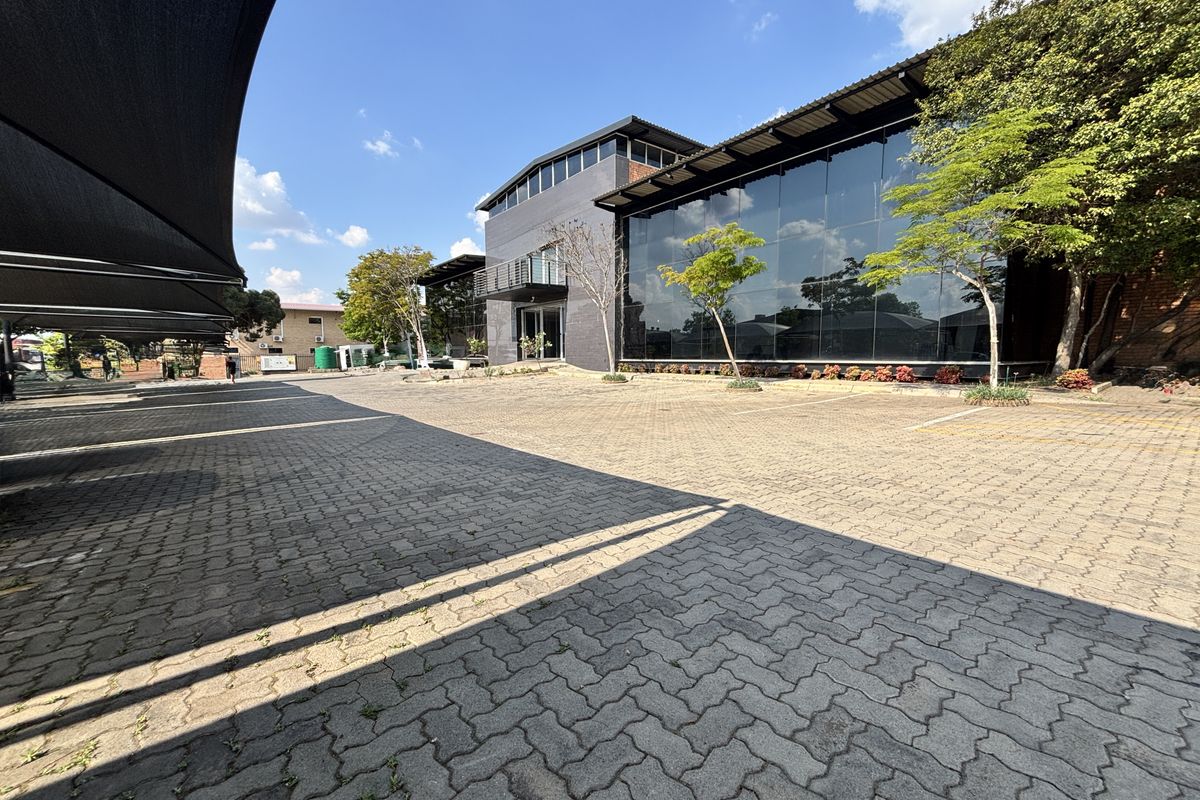 Modern industrial building featuring a large glass curtain wall facade, brick accents, and an extensive paved parking area with shade cloth carports.