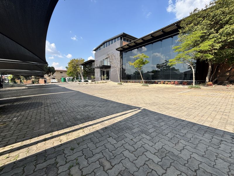 Modern industrial building featuring a large glass curtain wall facade, brick accents, and an extensive paved parking area with shade cloth carports.