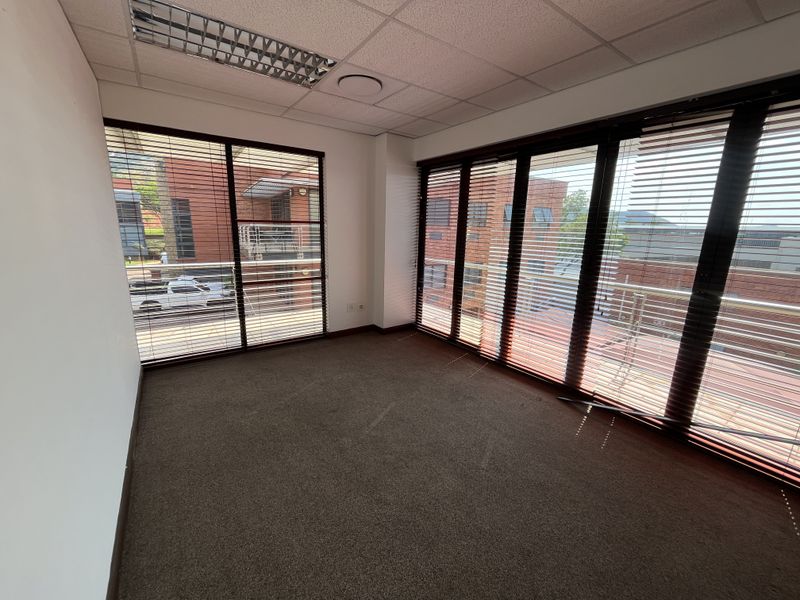 A bright, empty office space featuring carpeted flooring, a suspended ceiling with recessed lighting, and large glass doors providing ample natural light.