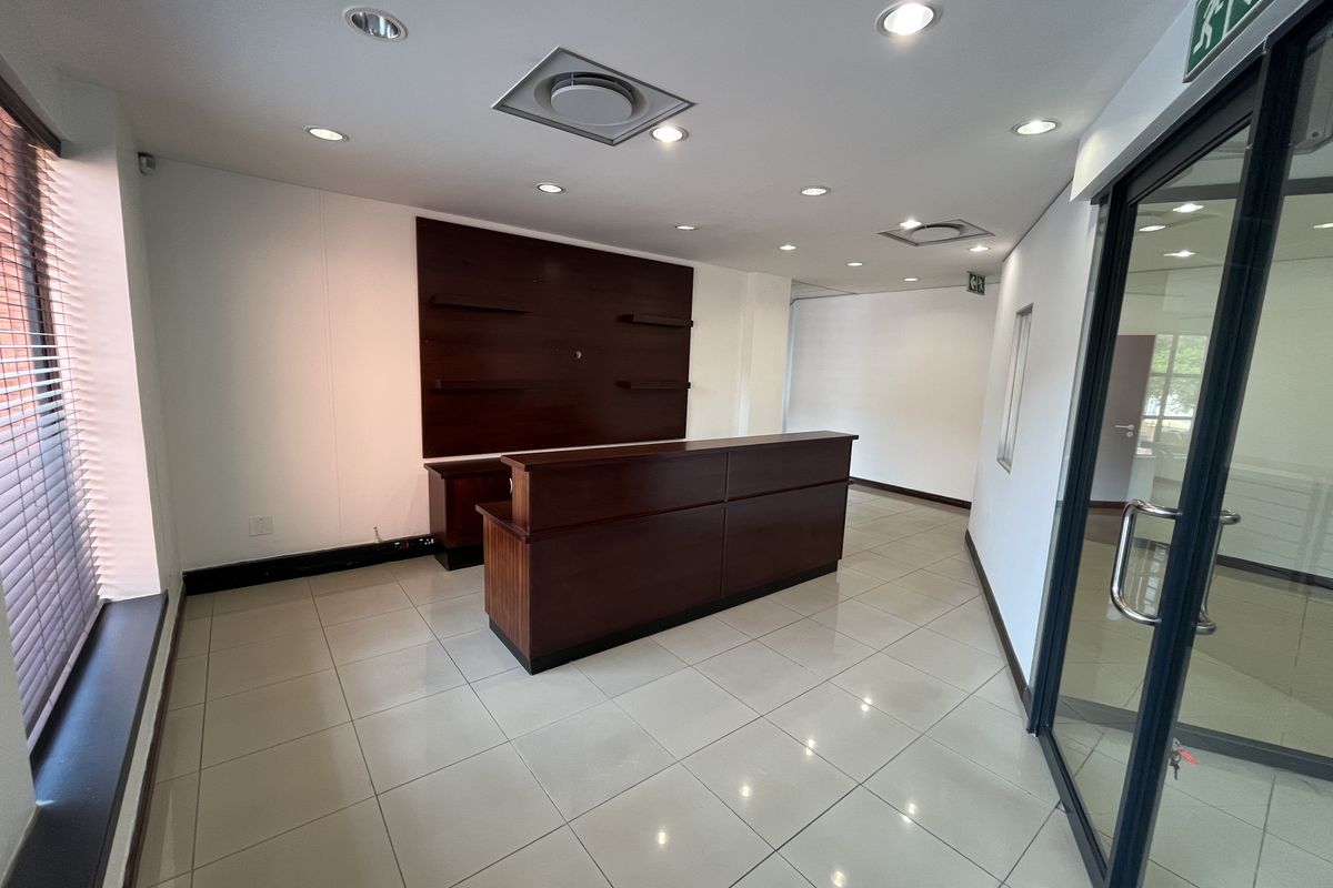 A professional office reception area featuring a built-in wooden desk, tiled flooring, recessed lighting, and a glass entrance door.