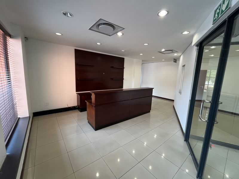 A professional office reception area featuring a built-in wooden desk, tiled flooring, recessed lighting, and a glass entrance door.
