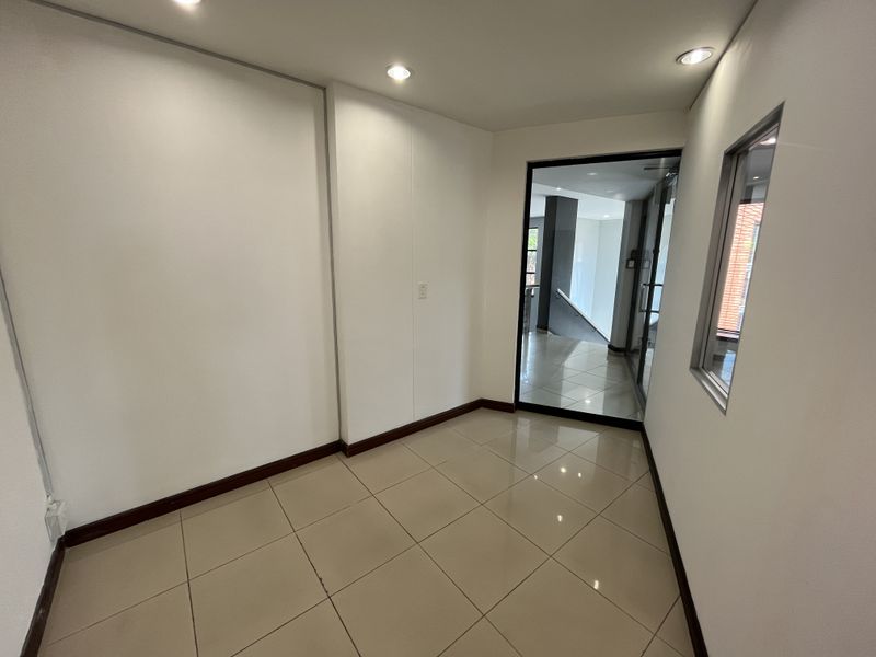 A clean, empty office space featuring polished porcelain floor tiles, recessed downlighting, and a modern glass partition door leading to the hallway.