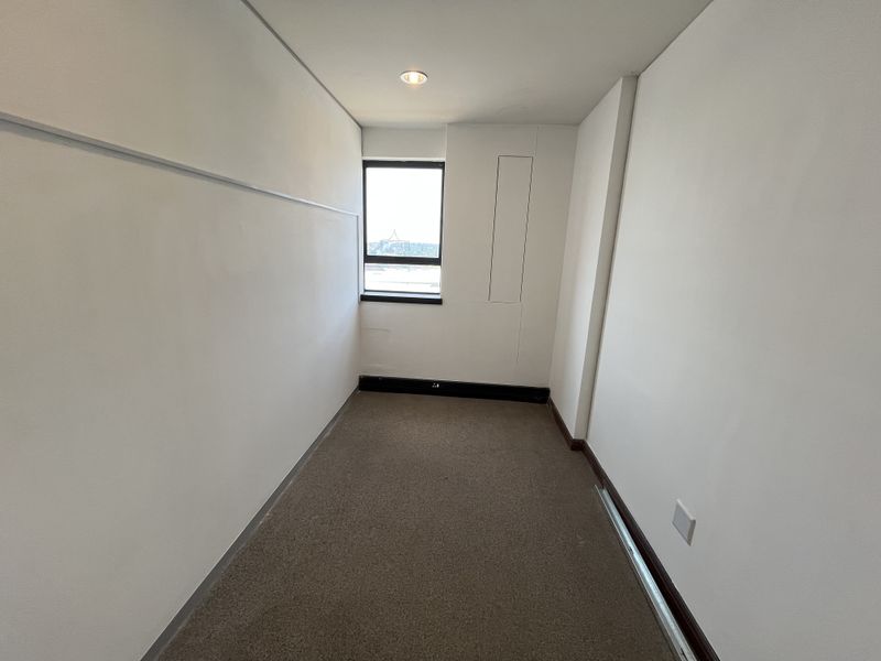 A compact, empty office space featuring neutral walls, carpeted flooring, and a single window providing natural light.