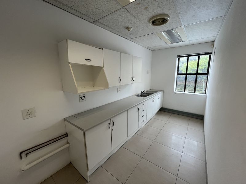 A functional office kitchenette featuring built-in white cabinetry, a stainless steel sink, tiled flooring, and a drop ceiling with integrated lighting.