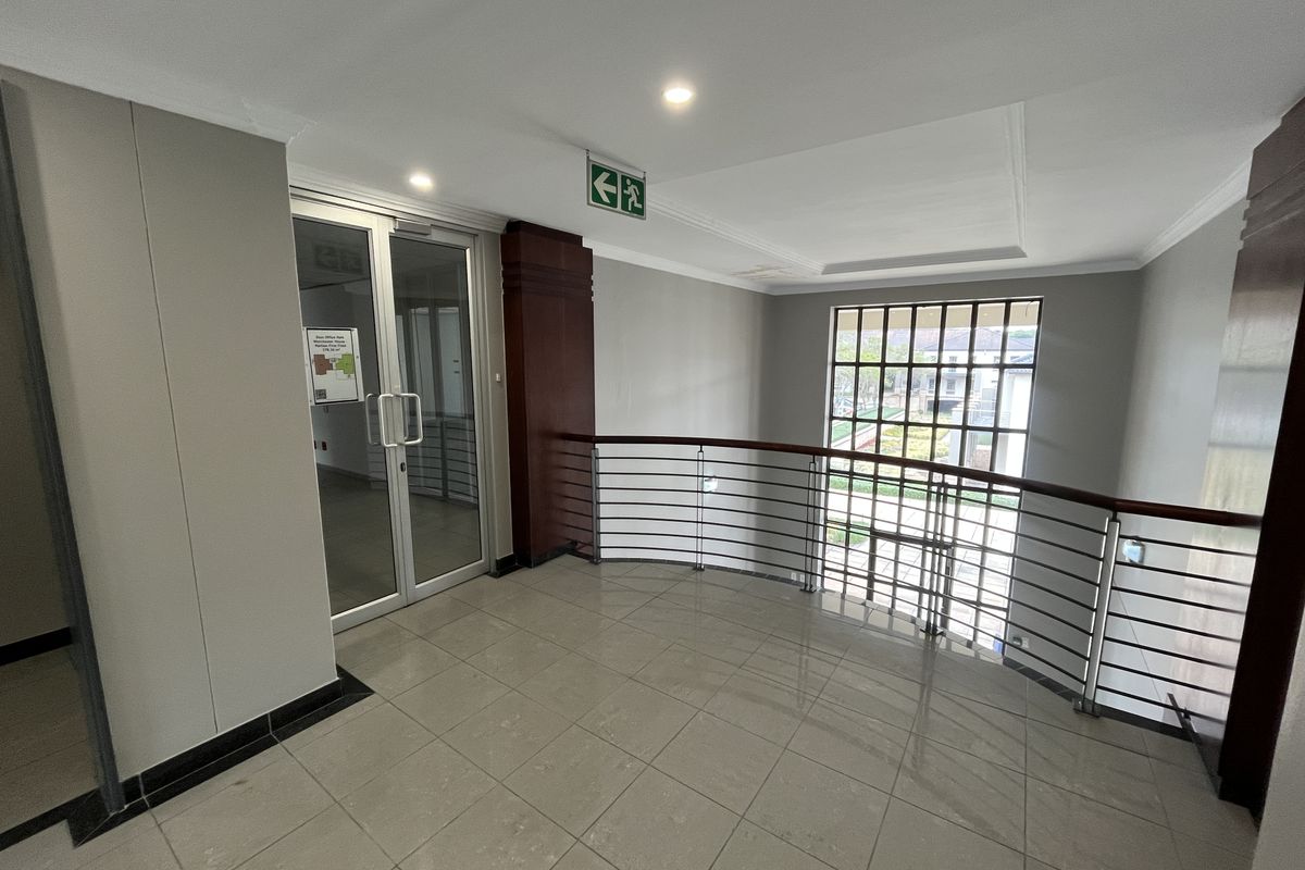 A bright, modern office hallway featuring tiled floors, a metal balustrade with a wooden handrail, and a glass entrance door.