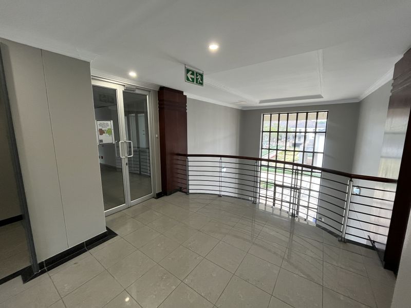 A bright, modern office hallway featuring tiled floors, a metal balustrade with a wooden handrail, and a glass entrance door.