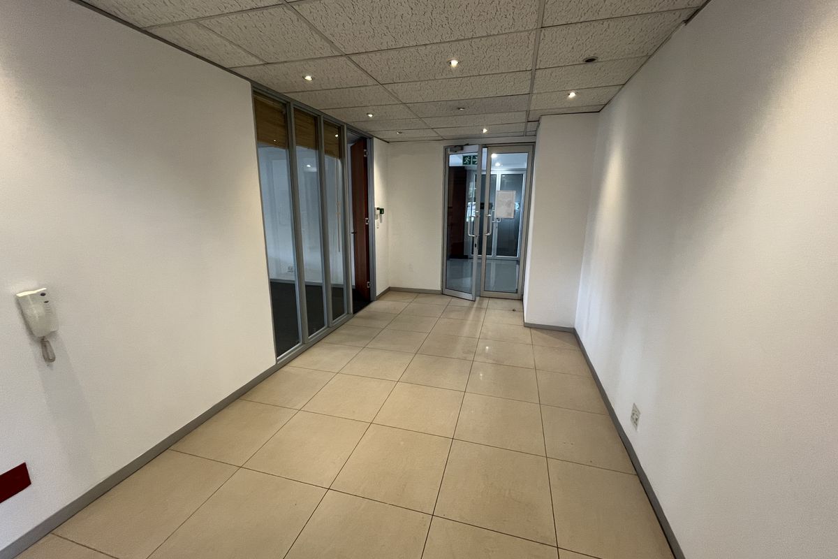 A clean, empty office hallway featuring tiled flooring, a suspended ceiling with recessed lighting, and glass partition doors leading to adjacent office spaces.