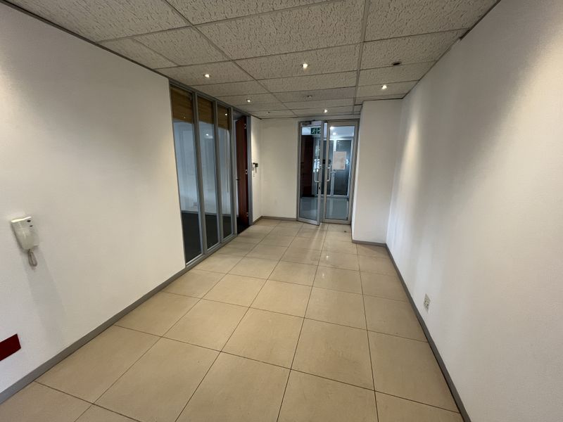 A clean, empty office hallway featuring tiled flooring, a suspended ceiling with recessed lighting, and glass partition doors leading to adjacent office spaces.
