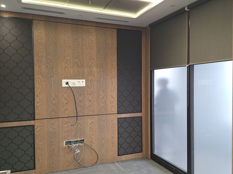 A modern office space featuring wood veneer wall paneling, patterned acoustic accents, recessed ceiling lighting, and carpet flooring, currently vacant with exposed wiring.