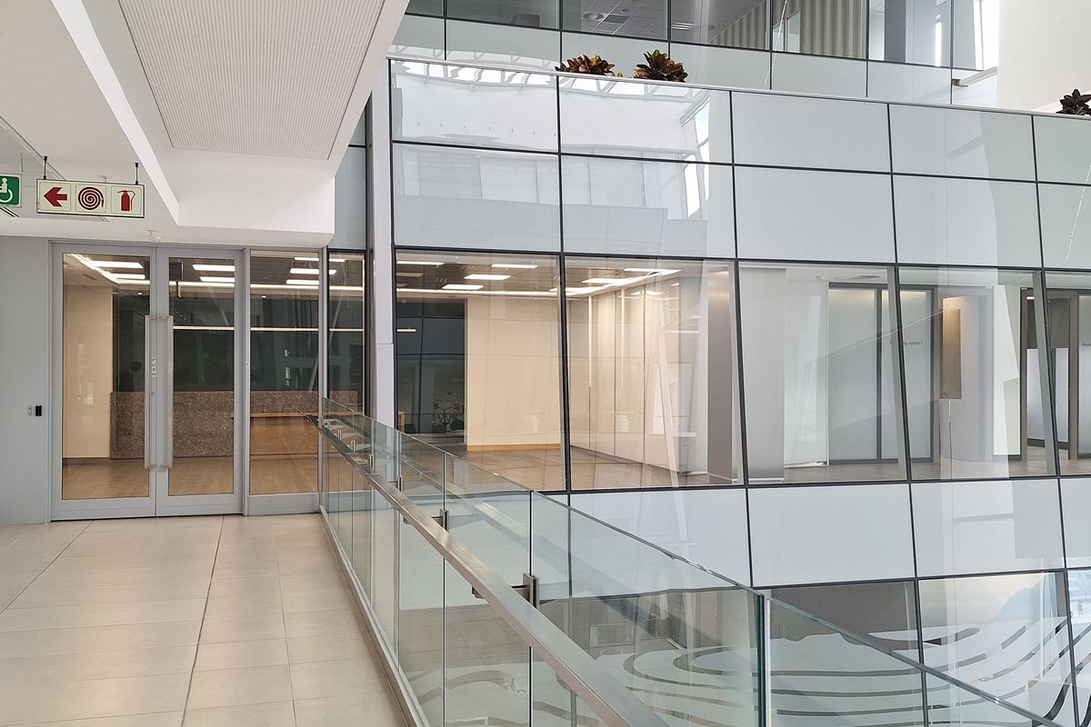 Modern office building interior featuring a spacious hallway with glass balustrades, tiled flooring, and extensive glass partitioning overlooking the atrium.