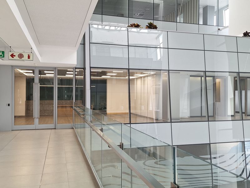 Modern office building interior featuring a spacious hallway with glass balustrades, tiled flooring, and extensive glass partitioning overlooking the atrium.