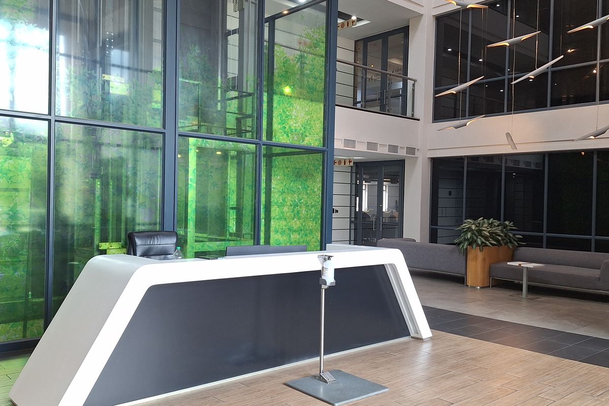 Modern office building reception area featuring a high-ceiling atrium, glass elevator shaft, and contemporary reception desk with tiled flooring.