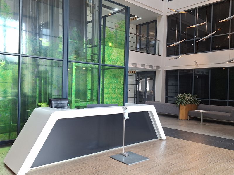 Modern office building reception area featuring a high-ceiling atrium, glass elevator shaft, and contemporary reception desk with tiled flooring.