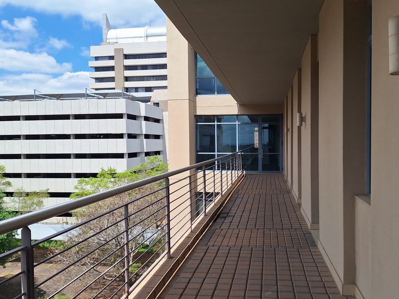 A clean, covered outdoor balcony featuring paved flooring and a modern stainless steel railing, providing access to the office building interior.