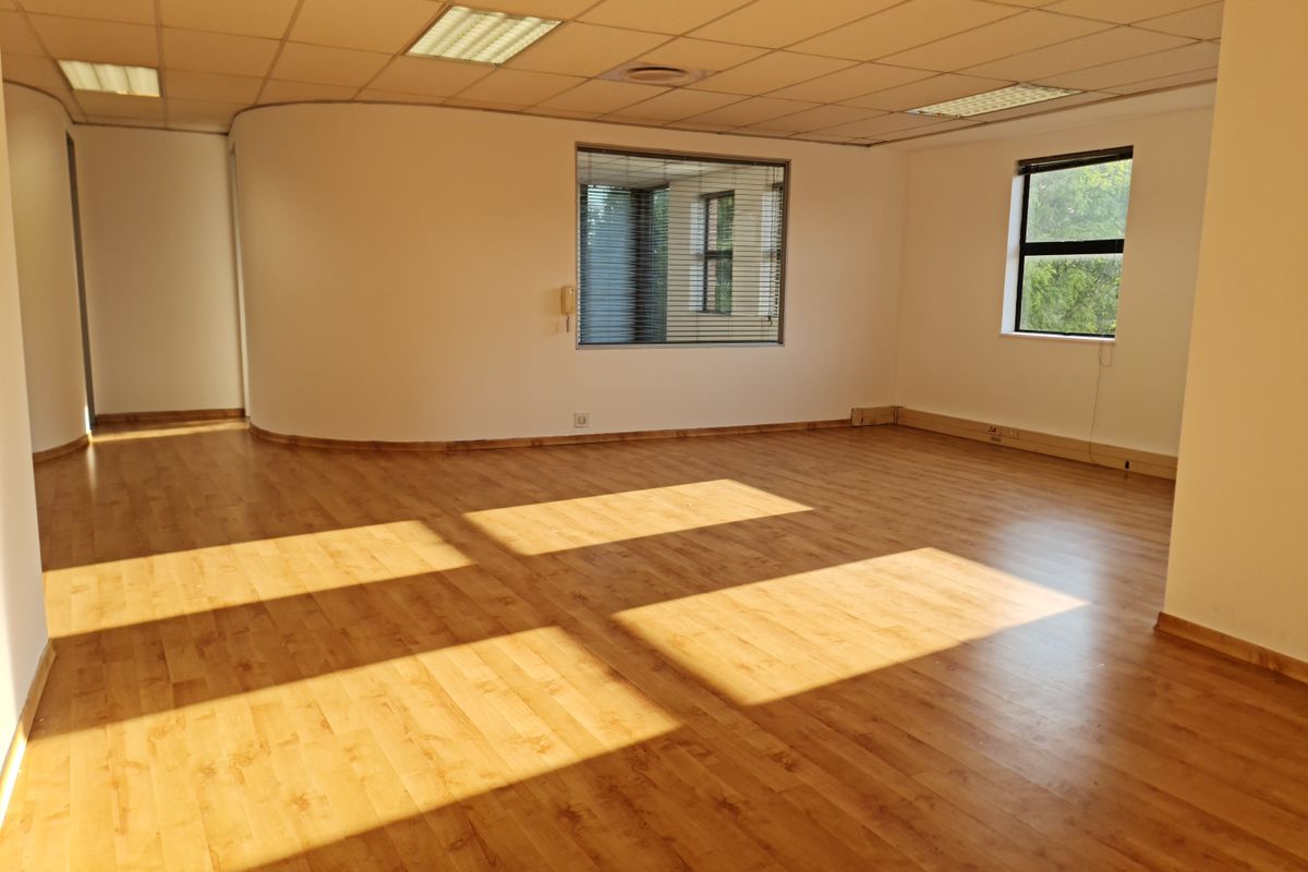 Spacious, empty office area featuring laminate flooring, a curved wall design, suspended ceiling with recessed lighting, and large windows with fitted blinds.