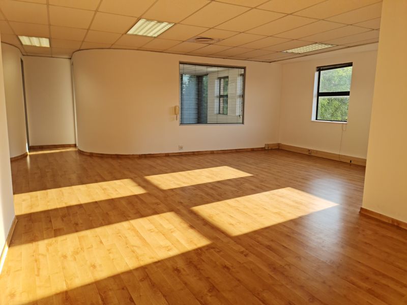 Spacious, empty office area featuring laminate flooring, a curved wall design, suspended ceiling with recessed lighting, and large windows with fitted blinds.
