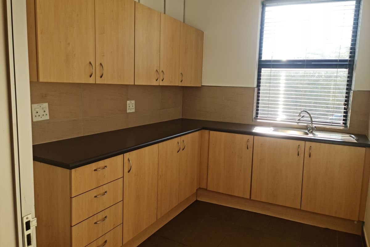A clean, functional office kitchenette featuring light wood-finish cabinetry, dark countertops, a stainless steel sink, and tiled flooring with natural light.