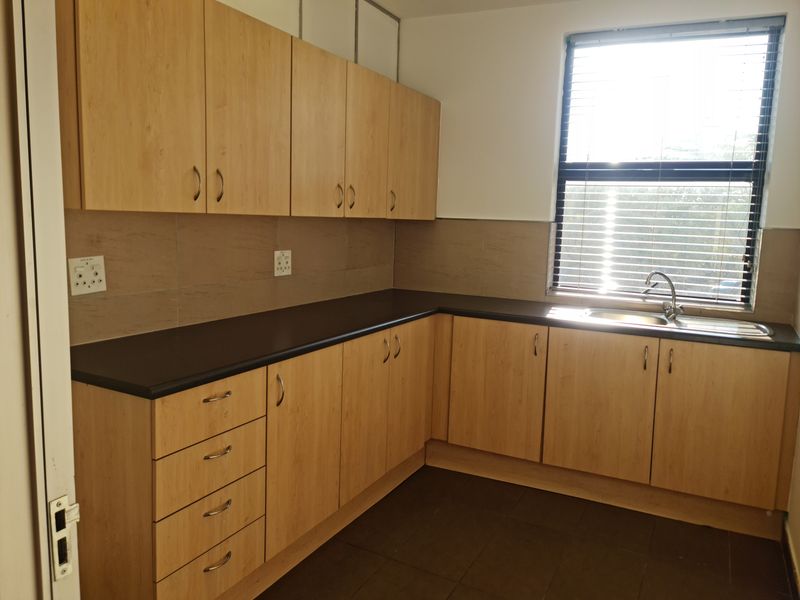 A clean, functional office kitchenette featuring light wood-finish cabinetry, dark countertops, a stainless steel sink, and tiled flooring with natural light.