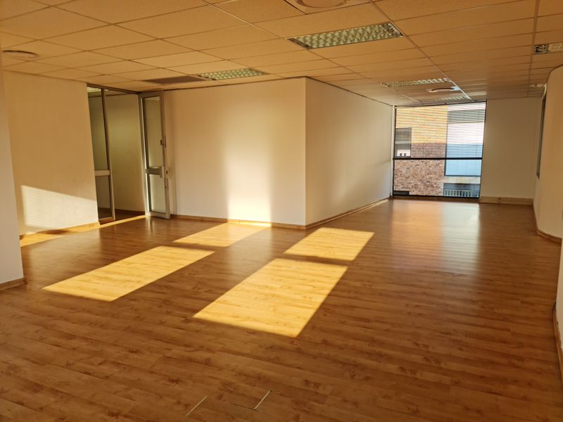 A spacious, empty office area featuring laminate flooring, a suspended ceiling with recessed lighting, and large windows providing natural light.