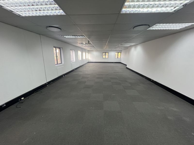 A spacious, empty open-plan office area featuring a suspended ceiling with recessed lighting, carpeted flooring, and multiple windows providing natural light.