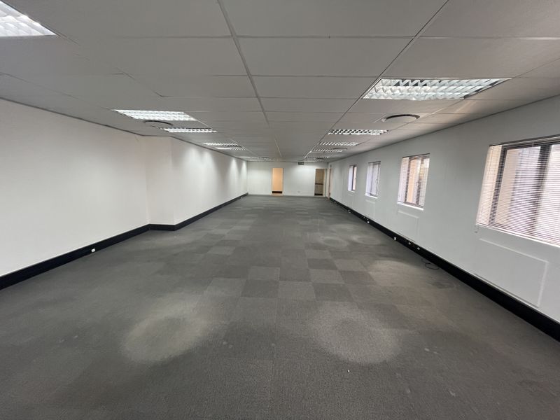 A spacious, empty open-plan office area featuring suspended ceilings, recessed lighting, and carpeted flooring with large windows and venetian blinds.