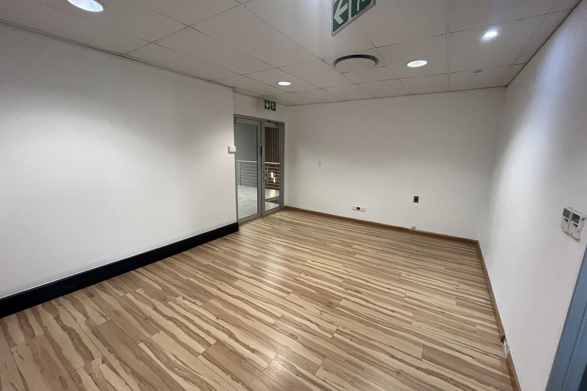A clean, empty office space featuring modern laminate flooring, a suspended ceiling with recessed lighting, and a glass door entrance.