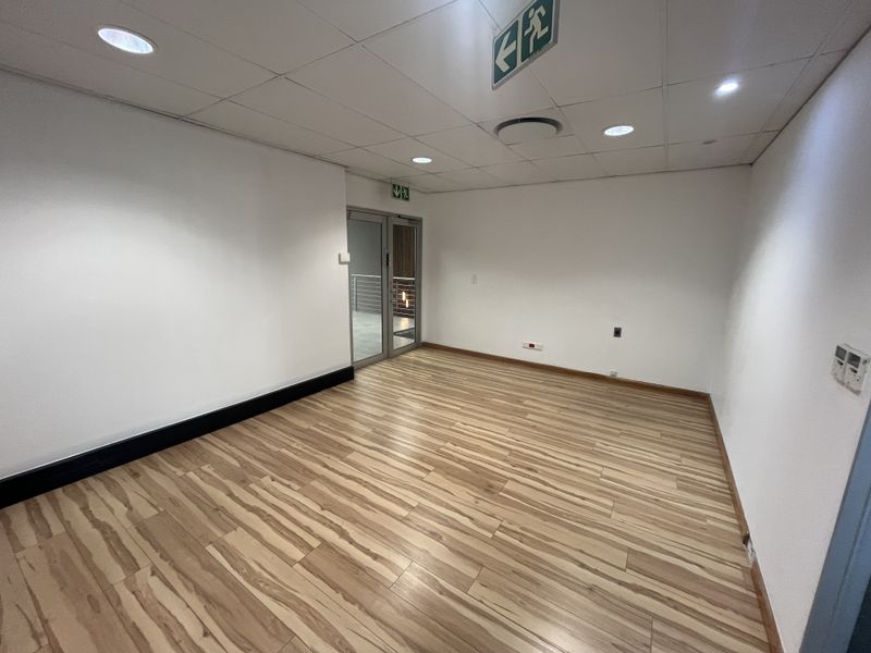 A clean, empty office space featuring modern laminate flooring, a suspended ceiling with recessed lighting, and a glass door entrance.