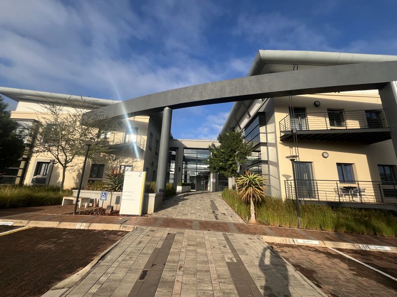 Modern office building exterior featuring a paved entrance, architectural archway, glass facade, and metal balconies under a clear sky.