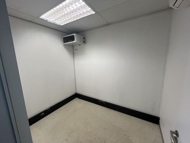 A compact, empty office space featuring a suspended ceiling, recessed lighting, wall-mounted server cabinet, and practical vinyl flooring with integrated power trunking.