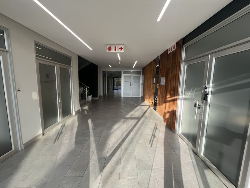 Modern office building hallway featuring tiled flooring, recessed linear lighting, timber wall paneling, and multiple glass-paneled office entrance doors.
