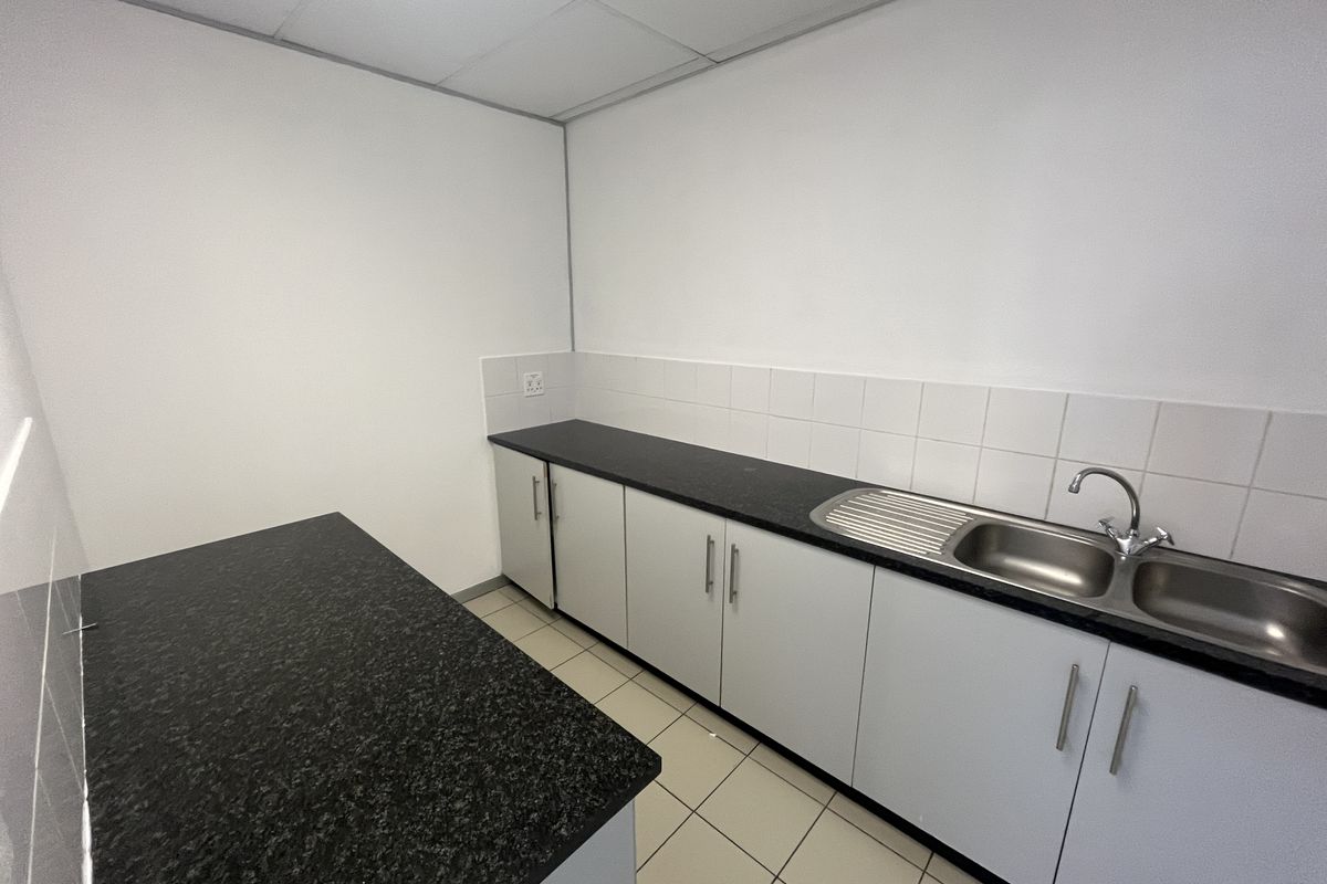 A clean, functional office kitchenette featuring white cabinetry, dark laminate countertops, a stainless steel double sink, and tiled flooring.