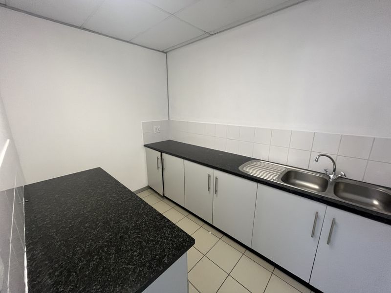 A clean, functional office kitchenette featuring white cabinetry, dark laminate countertops, a stainless steel double sink, and tiled flooring.