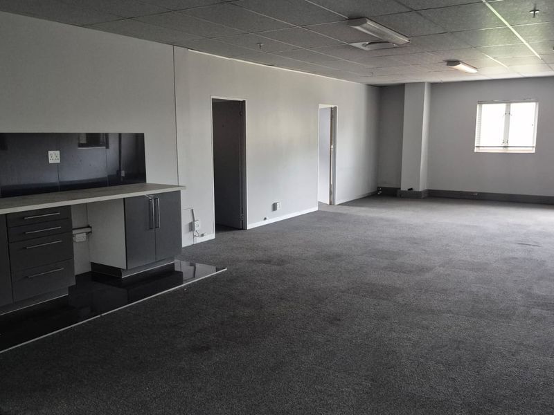 Spacious open-plan industrial office area featuring a kitchenette, carpeted flooring, drop ceiling, and large windows with venetian blinds.