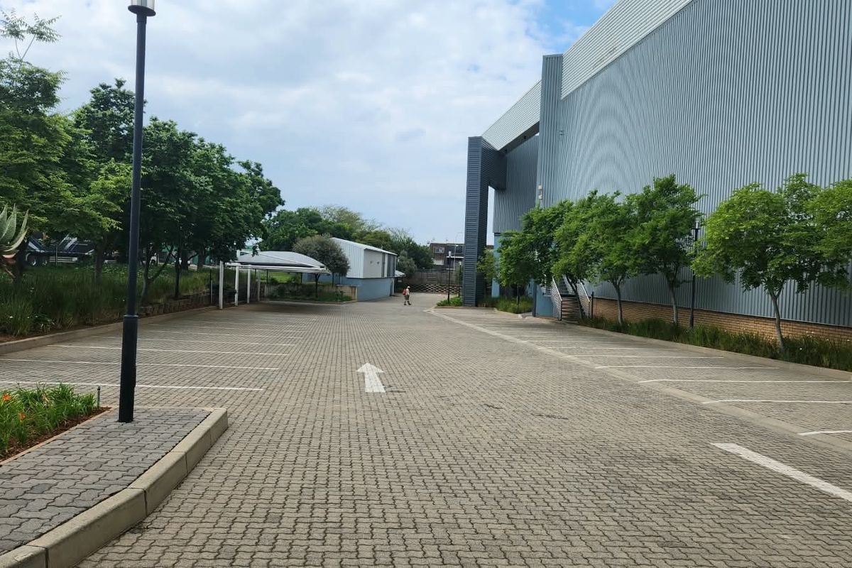 A spacious paved parking area adjacent to a large industrial building with metal cladding, featuring landscaped garden beds and a covered carport.