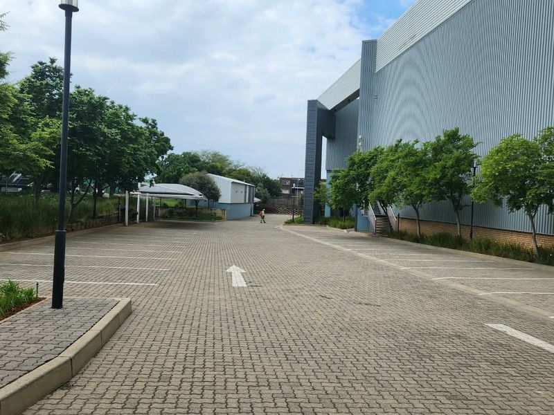 A spacious paved parking area adjacent to a large industrial building with metal cladding, featuring landscaped garden beds and a covered carport.