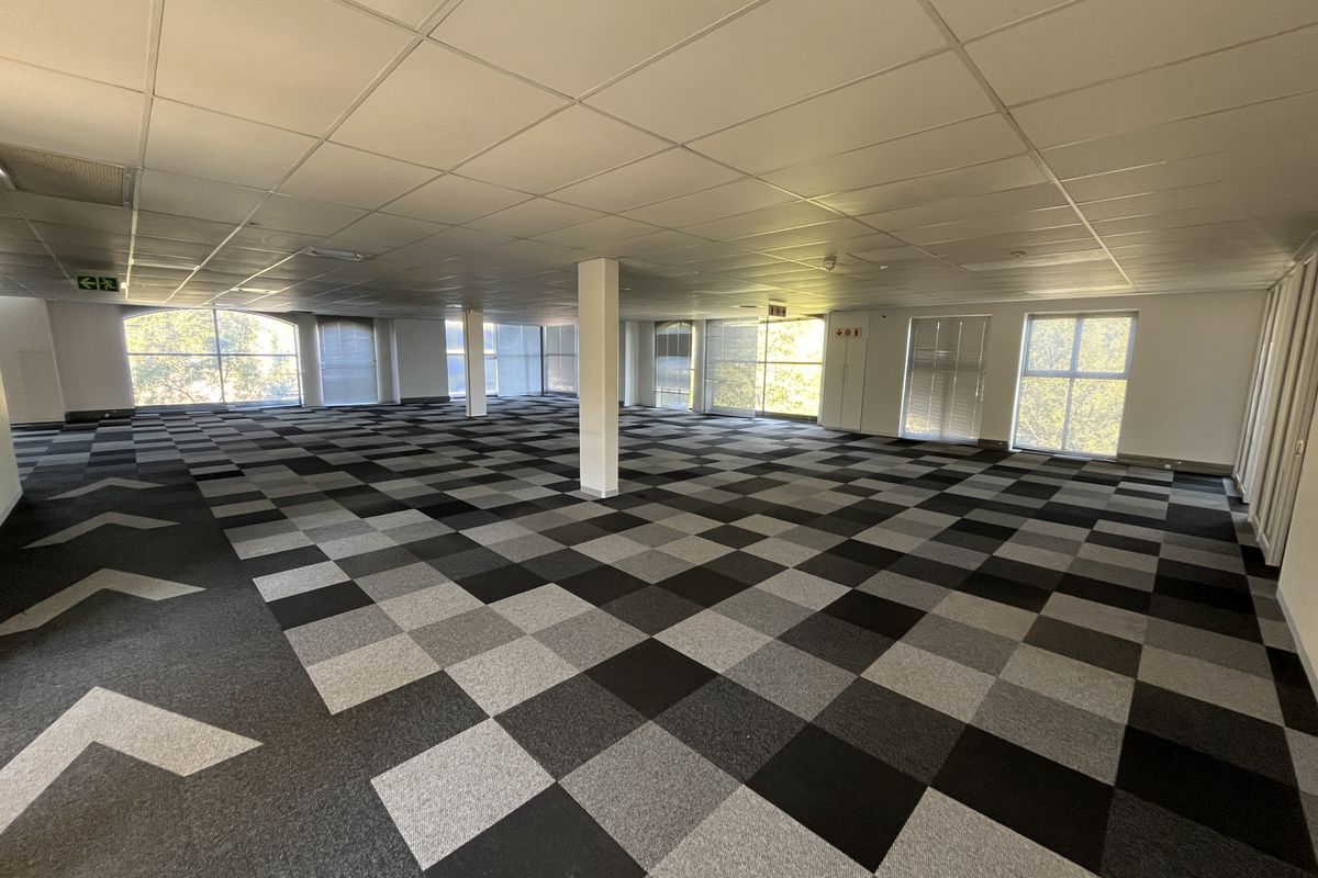 A spacious, open-plan office area featuring a suspended ceiling, patterned carpet tiles, and large windows providing ample natural light.