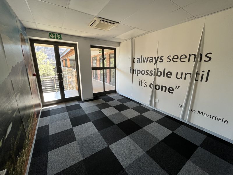 An empty office space featuring a suspended ceiling, recessed air conditioning, carpet tiles, and glass doors leading to an outdoor walkway.