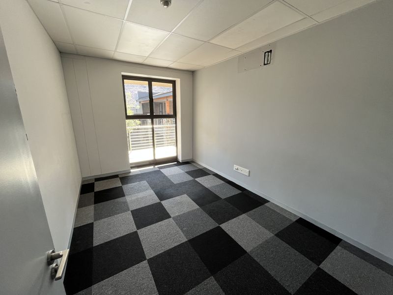 A vacant office space featuring a suspended ceiling, checkered carpet tiles, and a sliding glass door providing natural light.