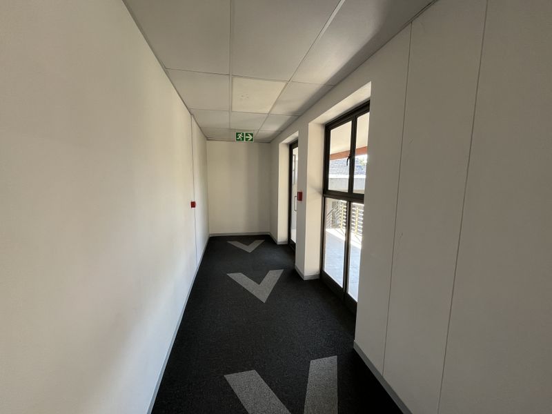 A clean, well-lit office hallway featuring a suspended ceiling, carpeted flooring with directional markings, and glass doors leading to an outdoor area.