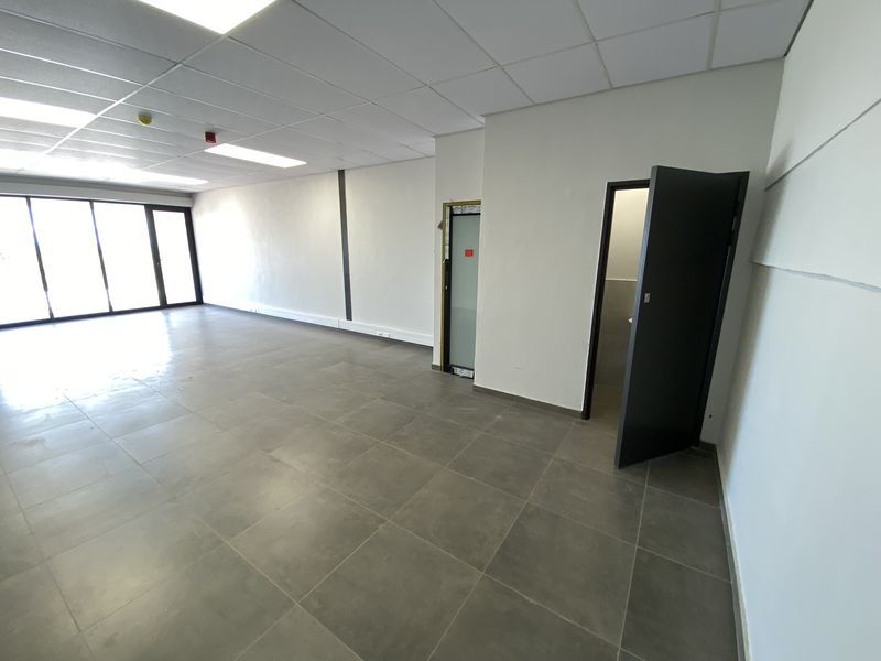 Open plan office space featuring tiled flooring, suspended ceiling with recessed lighting, and large glass doors providing natural light.