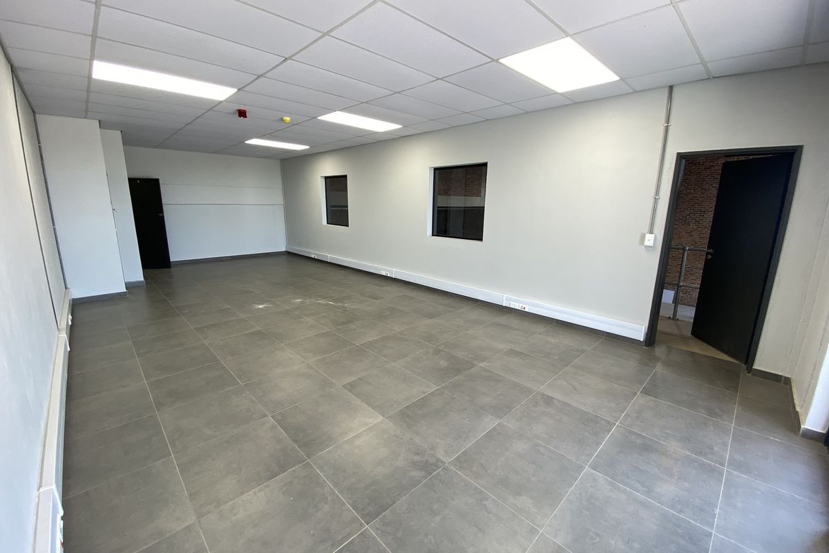 A spacious, empty industrial-style office featuring tiled flooring, a suspended ceiling with recessed lighting, and exposed brick accents near the entrance.